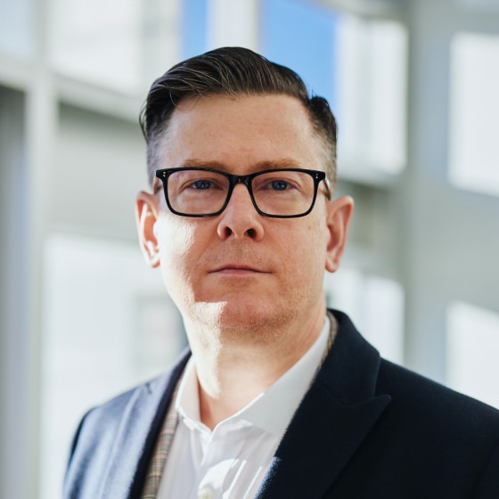 A person wearing glasses and a dark blazer stands indoors, with short hair, looking at the camera. Bright natural light filters through large windows in the background.