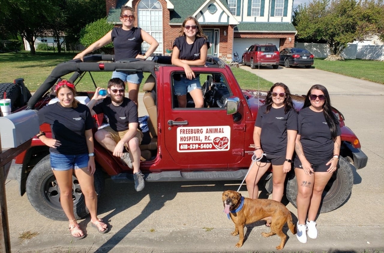 Seven people in black shirts and shorts, and a brown dog on a leash, pose around a red Jeep with a “Freeburg Animal Hospital” sign on a sunny suburban street.